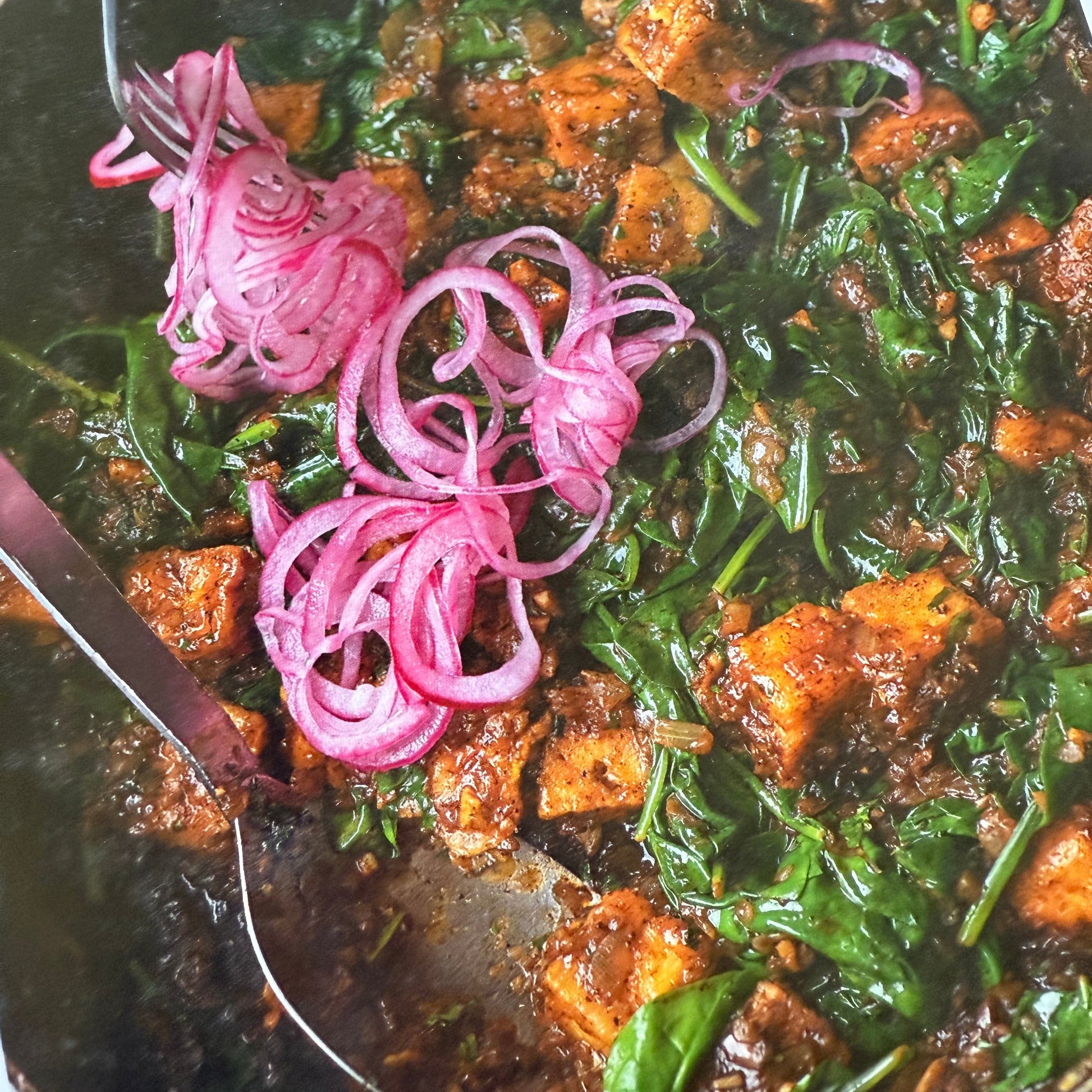 Sautéed greens with tofu and pink onions on a dark surface