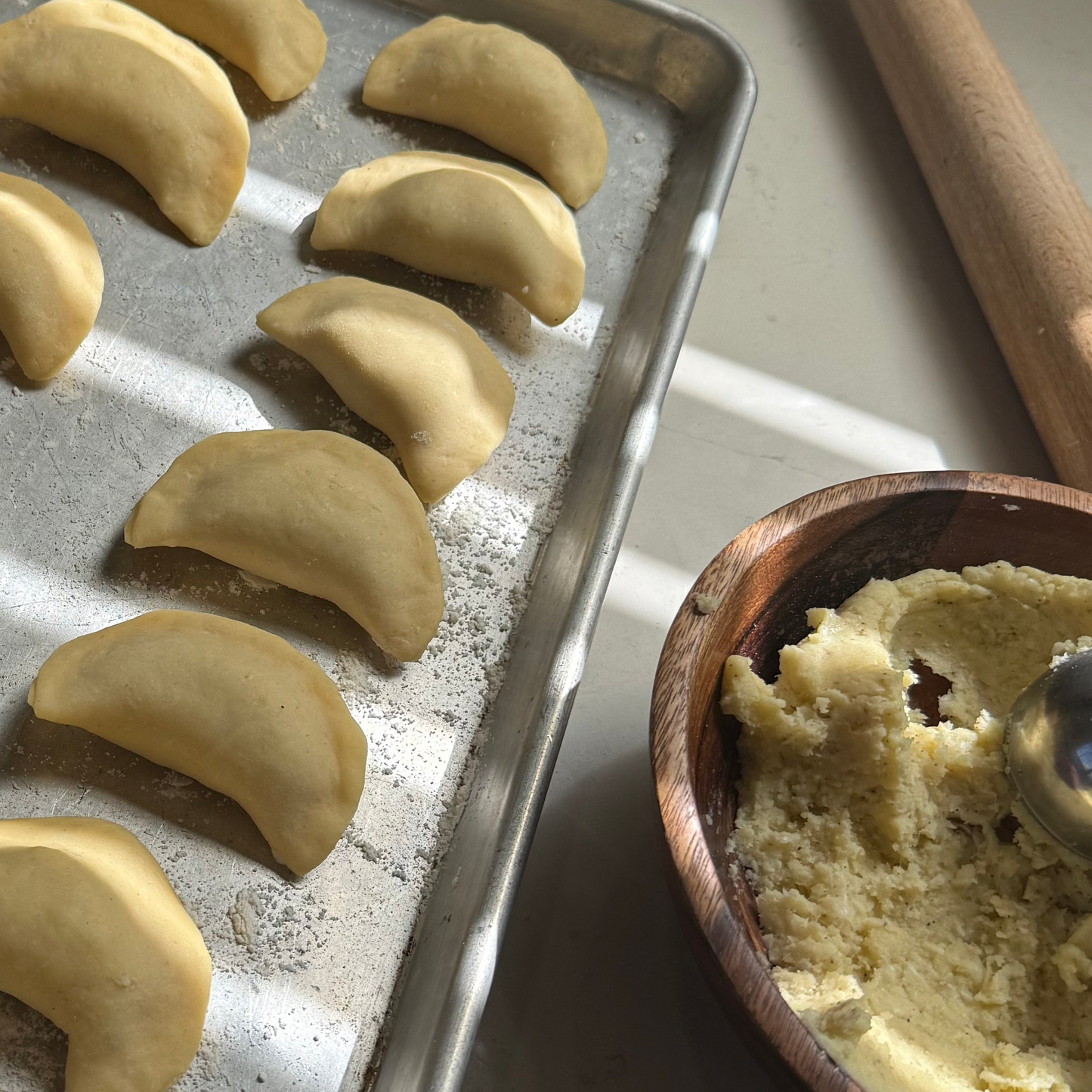 Uncooked dumplings on a baking sheet with a wooden bowl of dough and rolling pin.