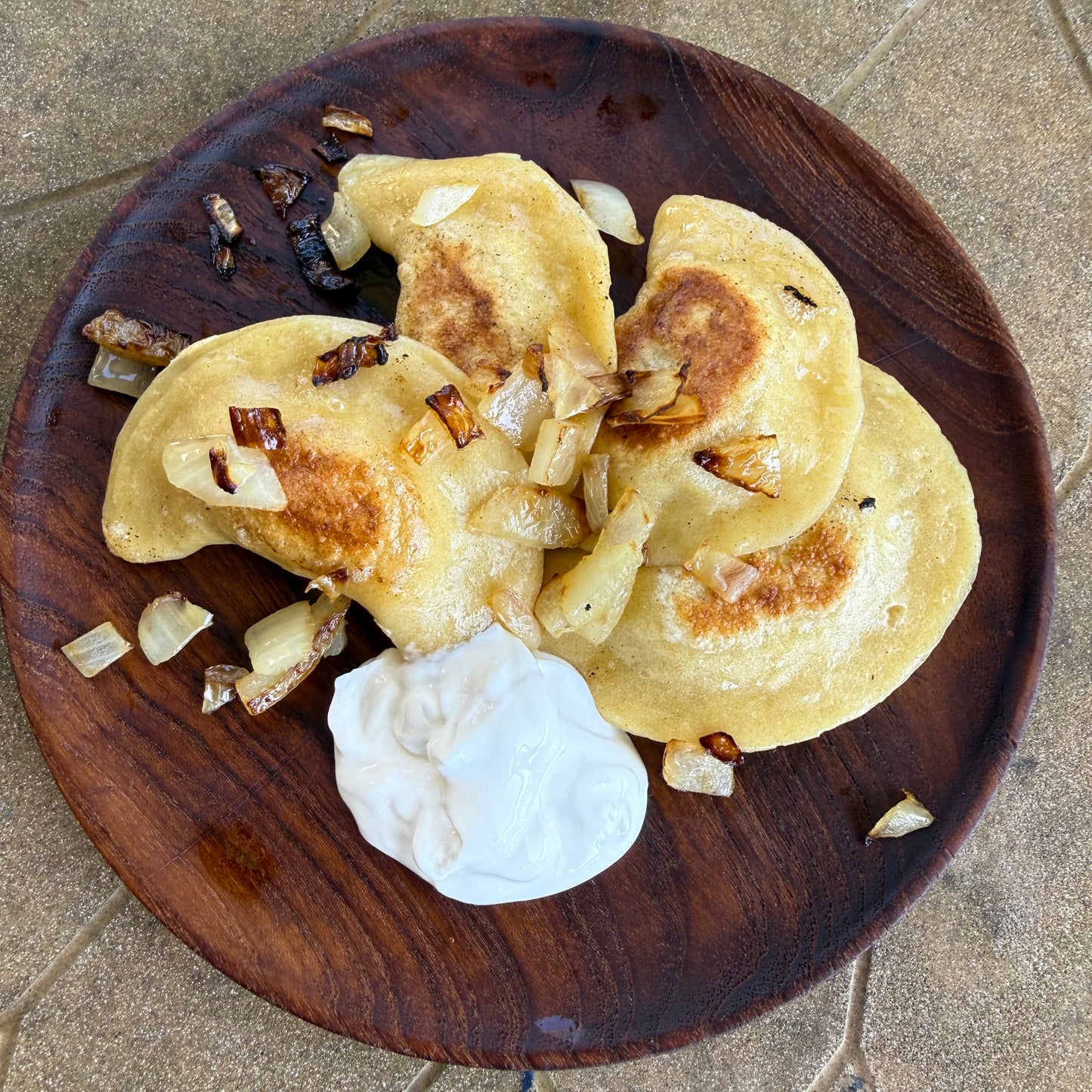 Three pierogies on a wooden plate with a dollop of sour cream on a stone surface.